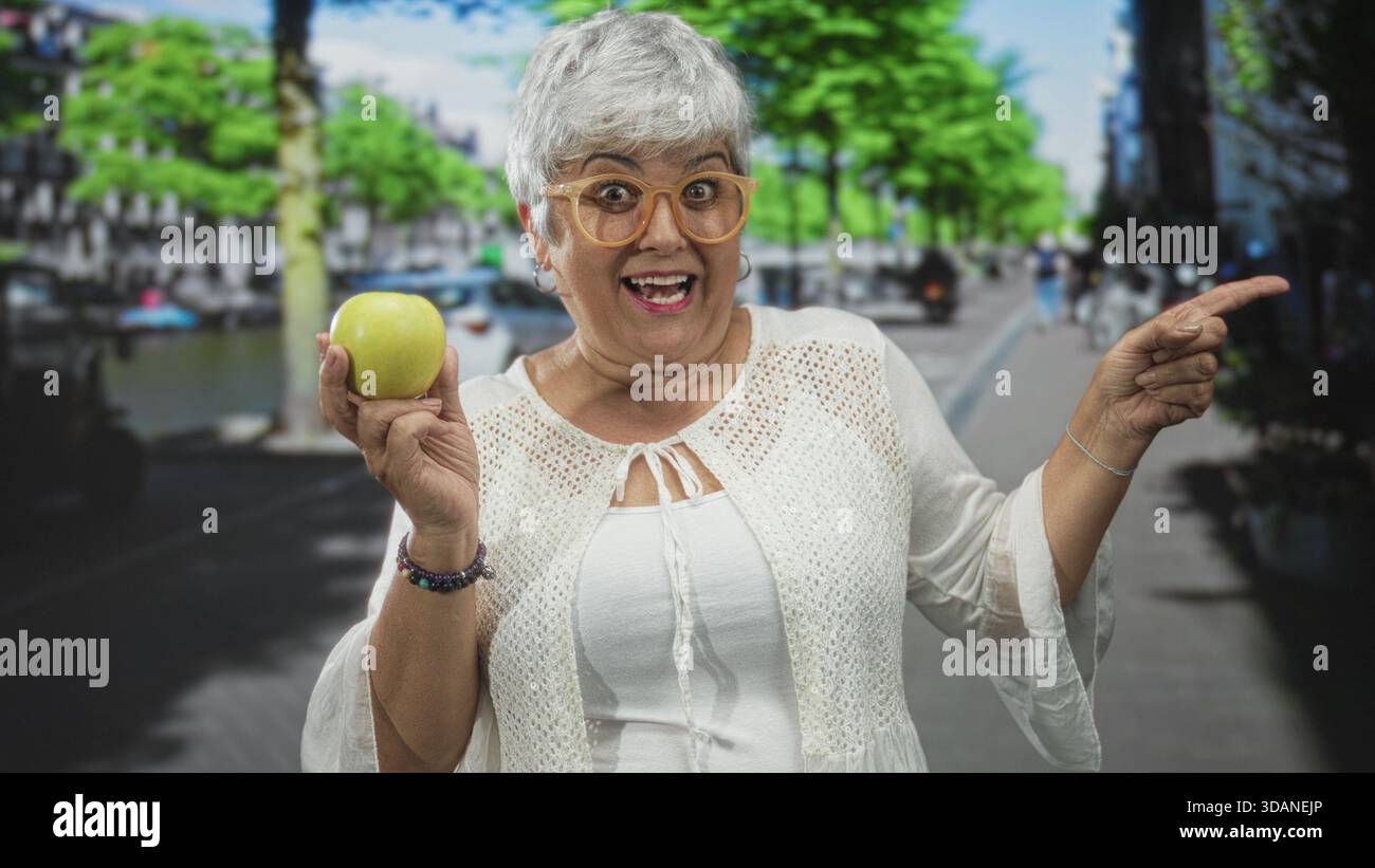 Donna che tiene la mela verde in mano sinistra e punta con la mano destra in strada, sorride con i capelli grigi e indossa gli occhiali; gioiosa curiosità. Foto Stock
