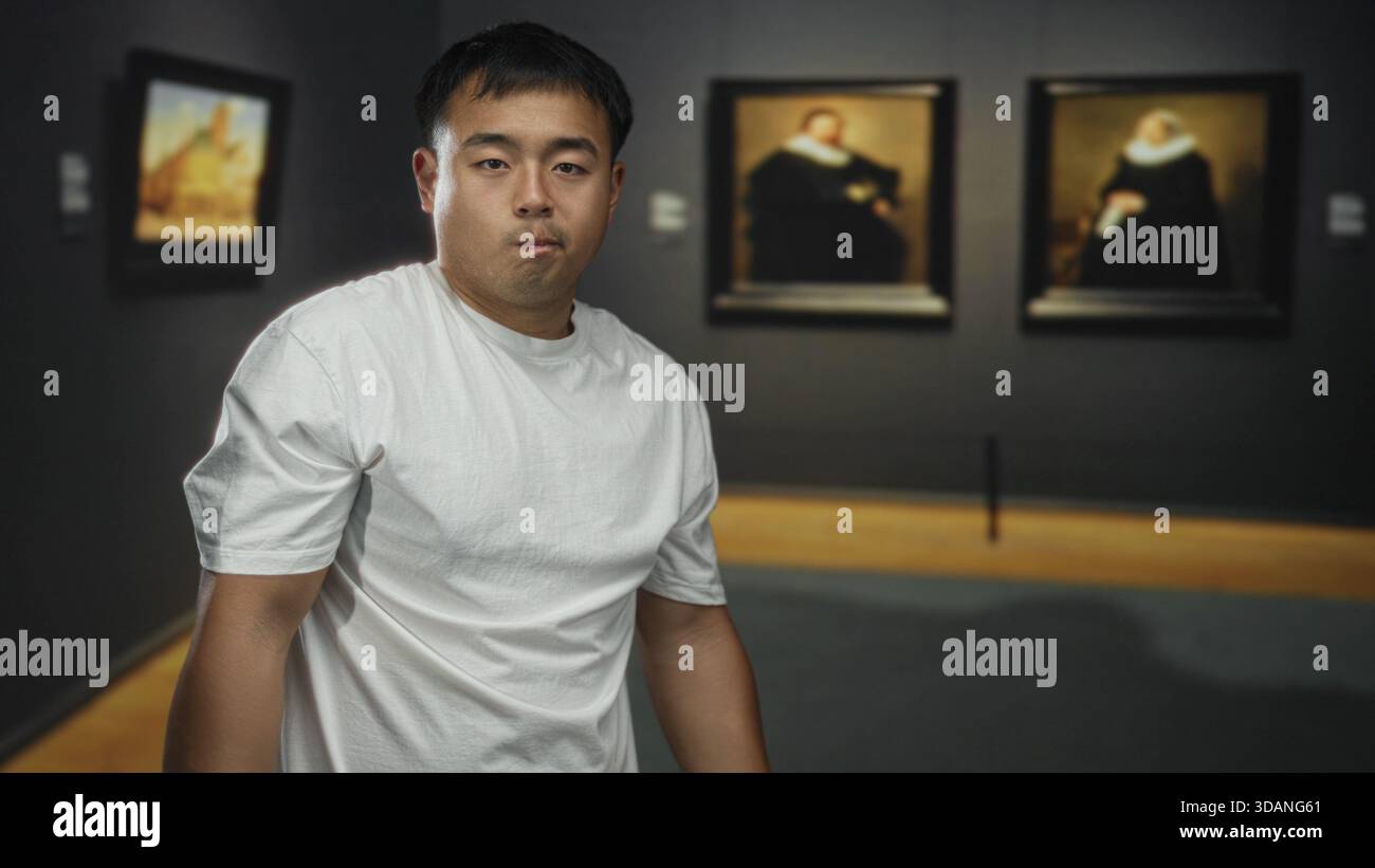 Il giovane cinese con la t-shirt bianca si ferma e fissa la pittura incorniciata sulle pareti in un edificio a galleria poco illuminato; contemplazione. Foto Stock