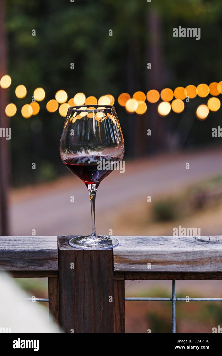 Un bicchiere di vino rosso poggia su una ringhiera di legno, adagiata su uno sfondo di soffici e scintillanti luci bokeh. Foto Stock
