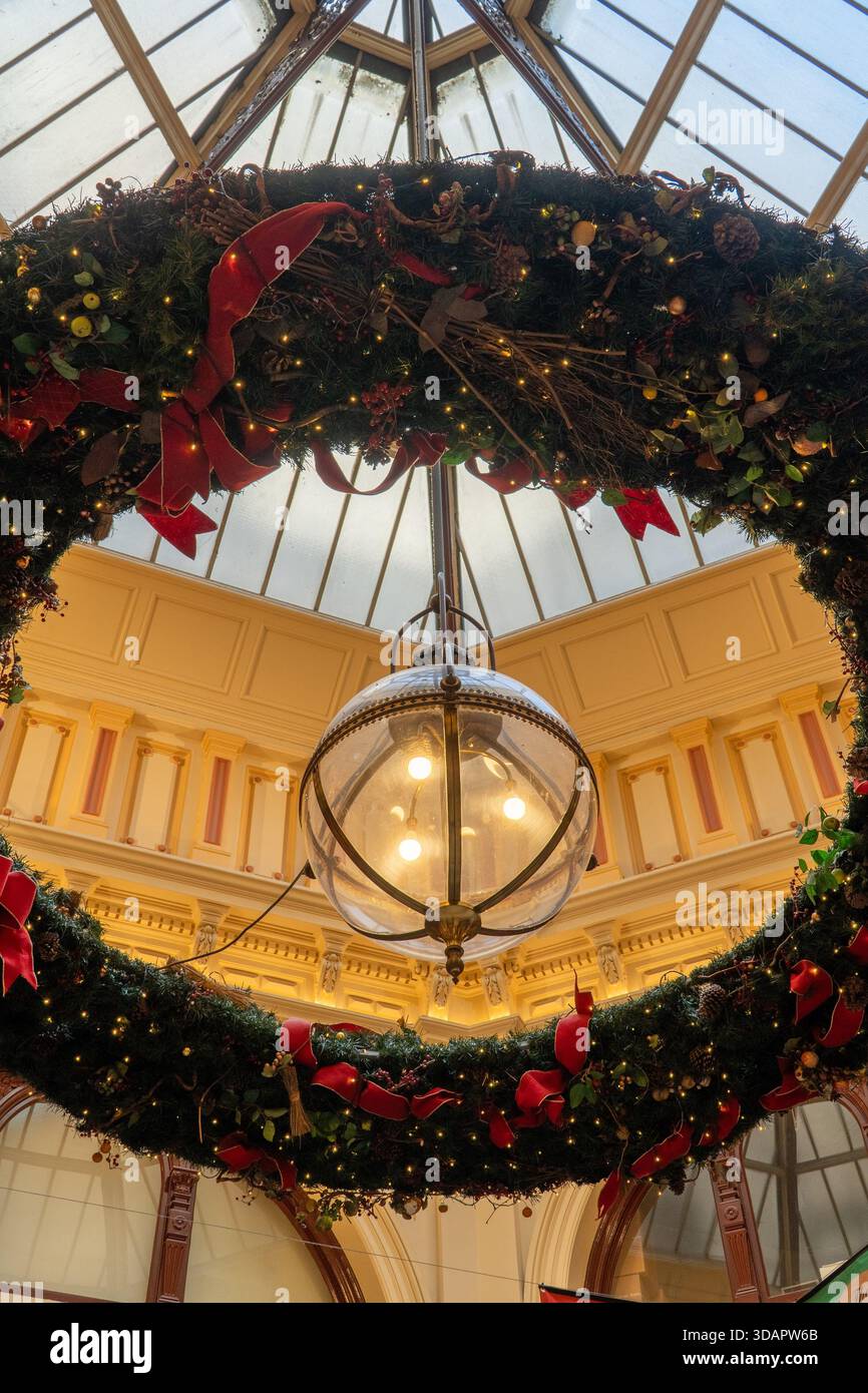 Una corona di Natale riccamente decorata, adornata con nastri rossi e luci festive, incornicia splendidamente un lampadario sferico luminoso sotto un ornato Foto Stock