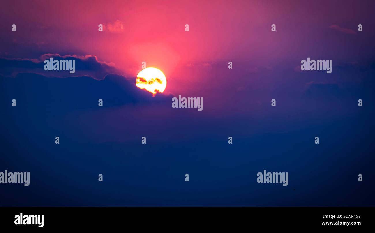 Tramonto vibrante con sole parzialmente oscurato dalle nuvole, creando una sfumatura dal rosa al blu profondo nel cielo. Foto Stock