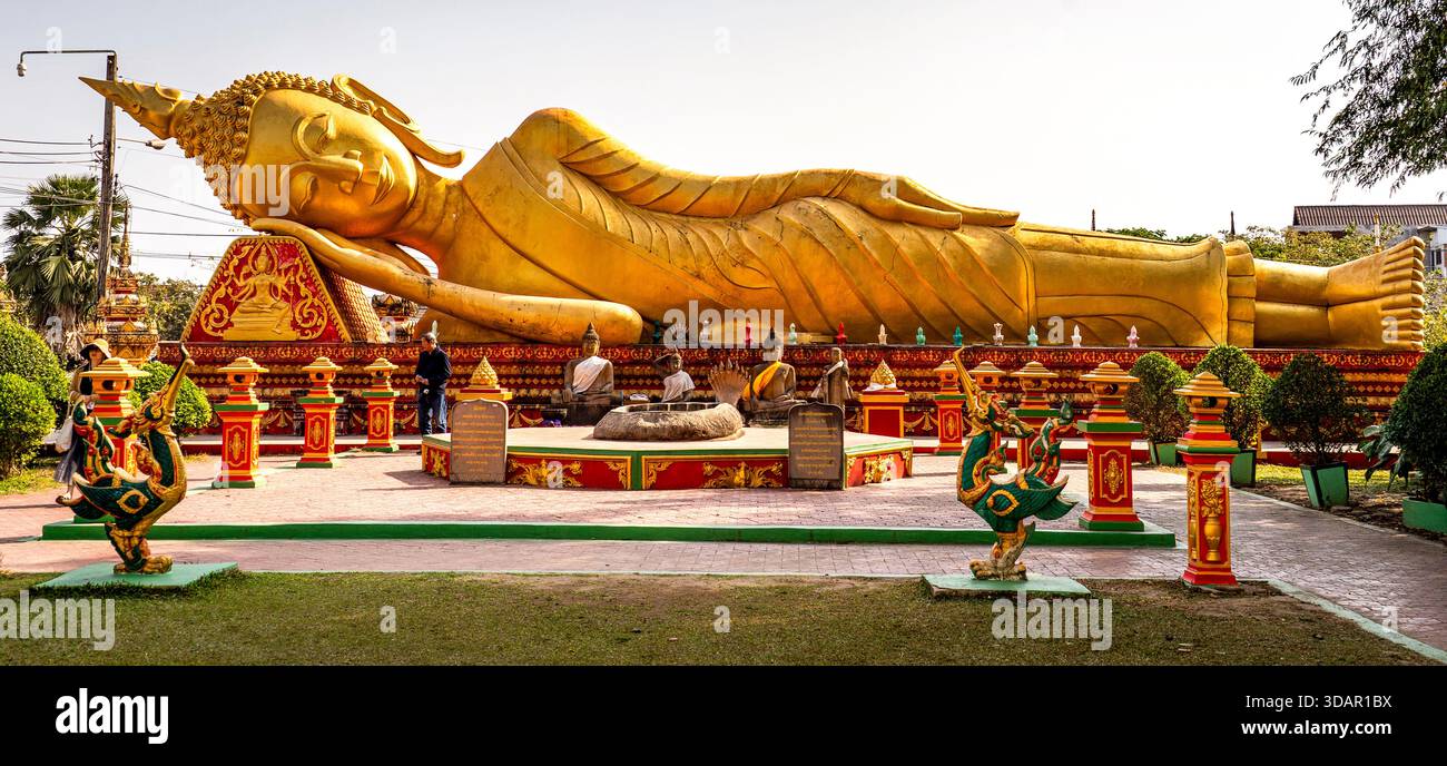 Statua dorata di Buddha reclinata in un giardino del tempio con decorazioni ornamentali e vegetazione, sotto un cielo limpido. Foto Stock