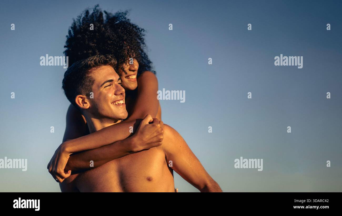 Una felice coppia interrazziale che si gode un giocoso giro in piggyback all'aperto durante l'ora d'oro, i loro volti illuminati da sorrisi radiosi contro un cielo blu limpido Foto Stock