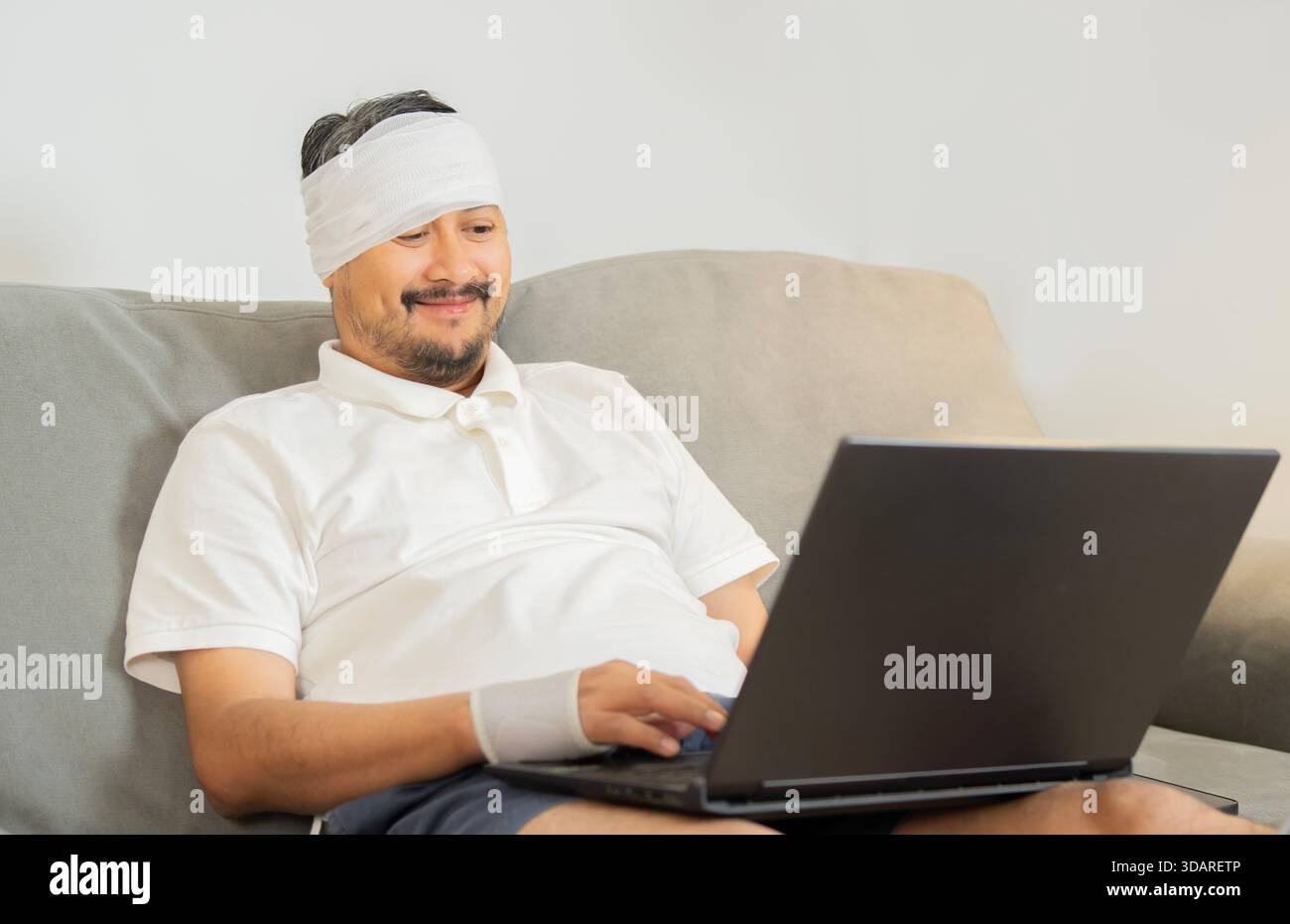 Bell'uomo allegro con un polso rotto e la testa in bendaggio seduto e che lavora a casa. Trauma e riabilitazione. Sicurezza sociale e salute insur Foto Stock