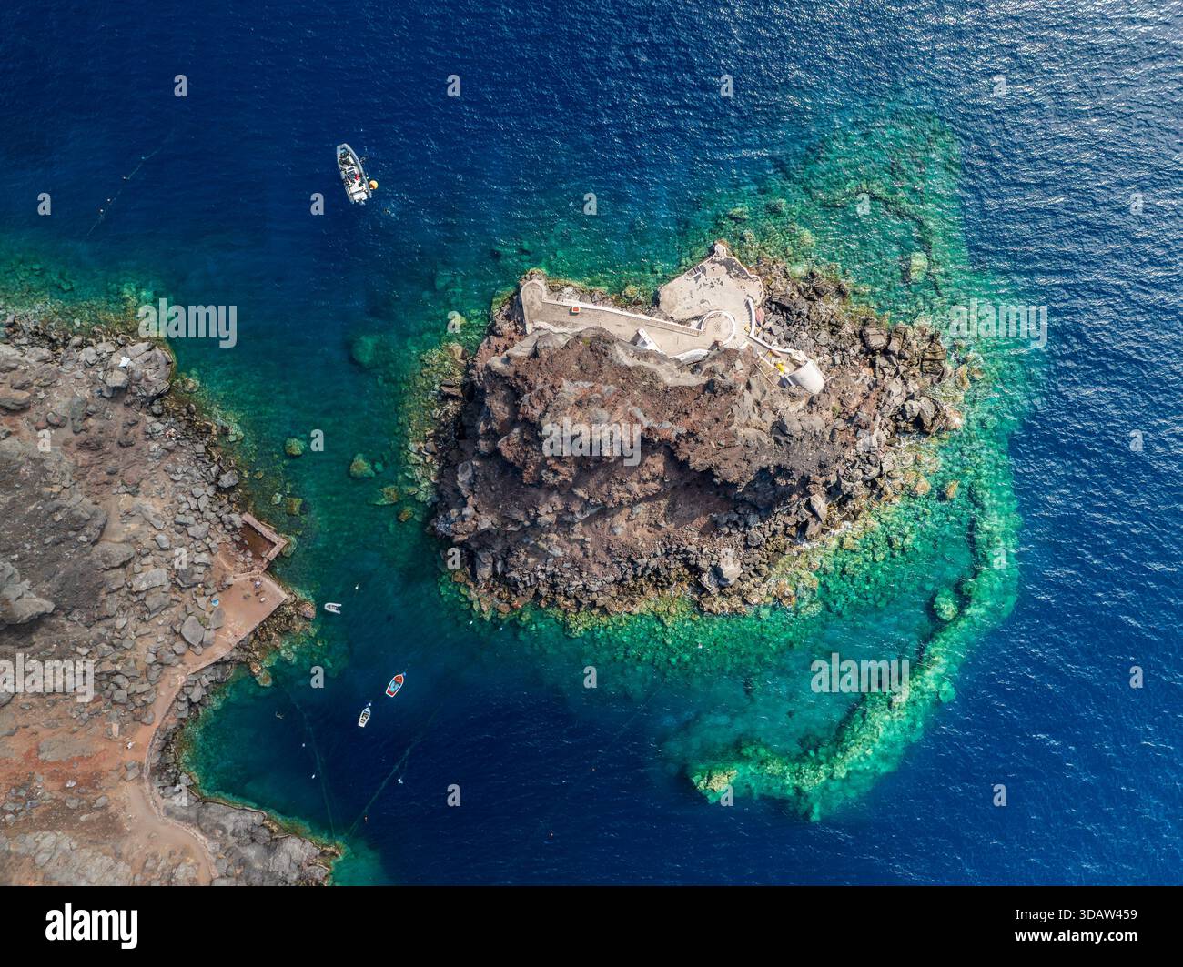 Vista aerea di un'aspra isola vulcanica circondata da acque turchesi, in contrasto con il mare blu profondo che la circonda, Oia, Thira, Grecia. Foto Stock