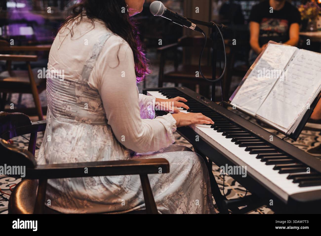Una donna corta e irriconoscibile si esibisce a un concerto, suona una tastiera elettronica e canta in un microfono, con spartiti su un piedistallo, creatina Foto Stock
