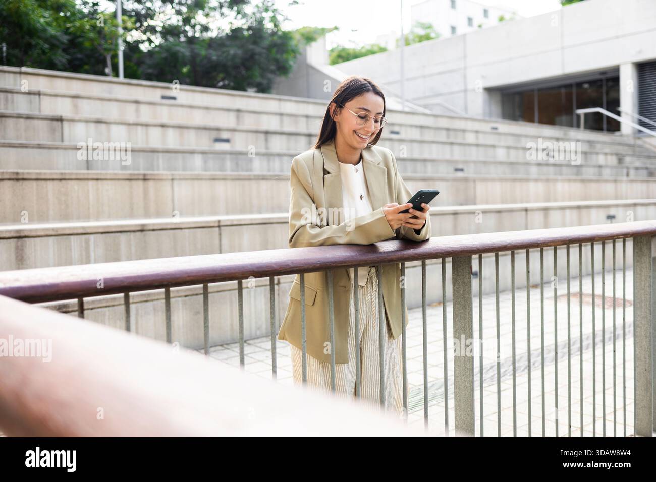 Una donna d'affari in piedi fuori, appoggiata su una ringhiera, mentre usa il suo smartphone. È sorridente, vestita con un abbigliamento professionale, e sembra impegnarsi Foto Stock