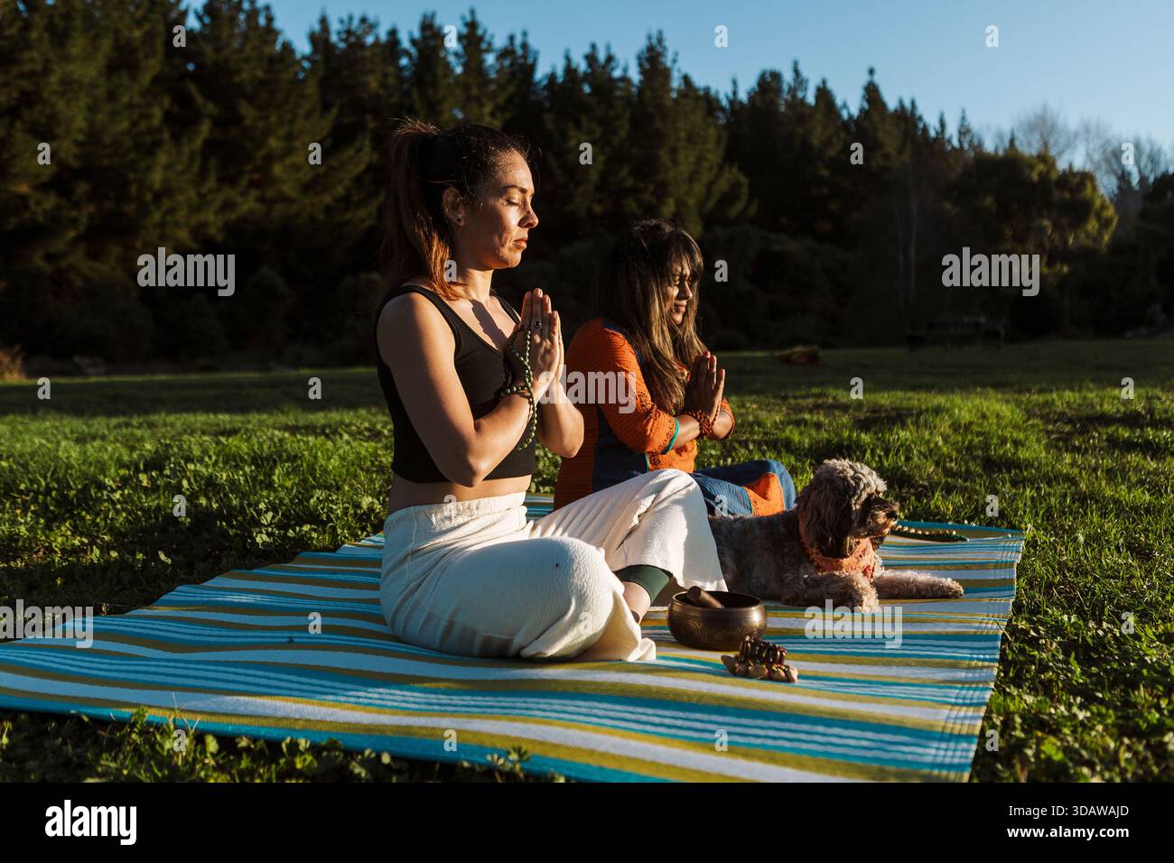 Due donne praticano il bagno all'aperto in nuova Zelanda durante l'autunno, sedute su un tappeto a righe circondato dalla natura, meditando e godendosi il tranqui Foto Stock