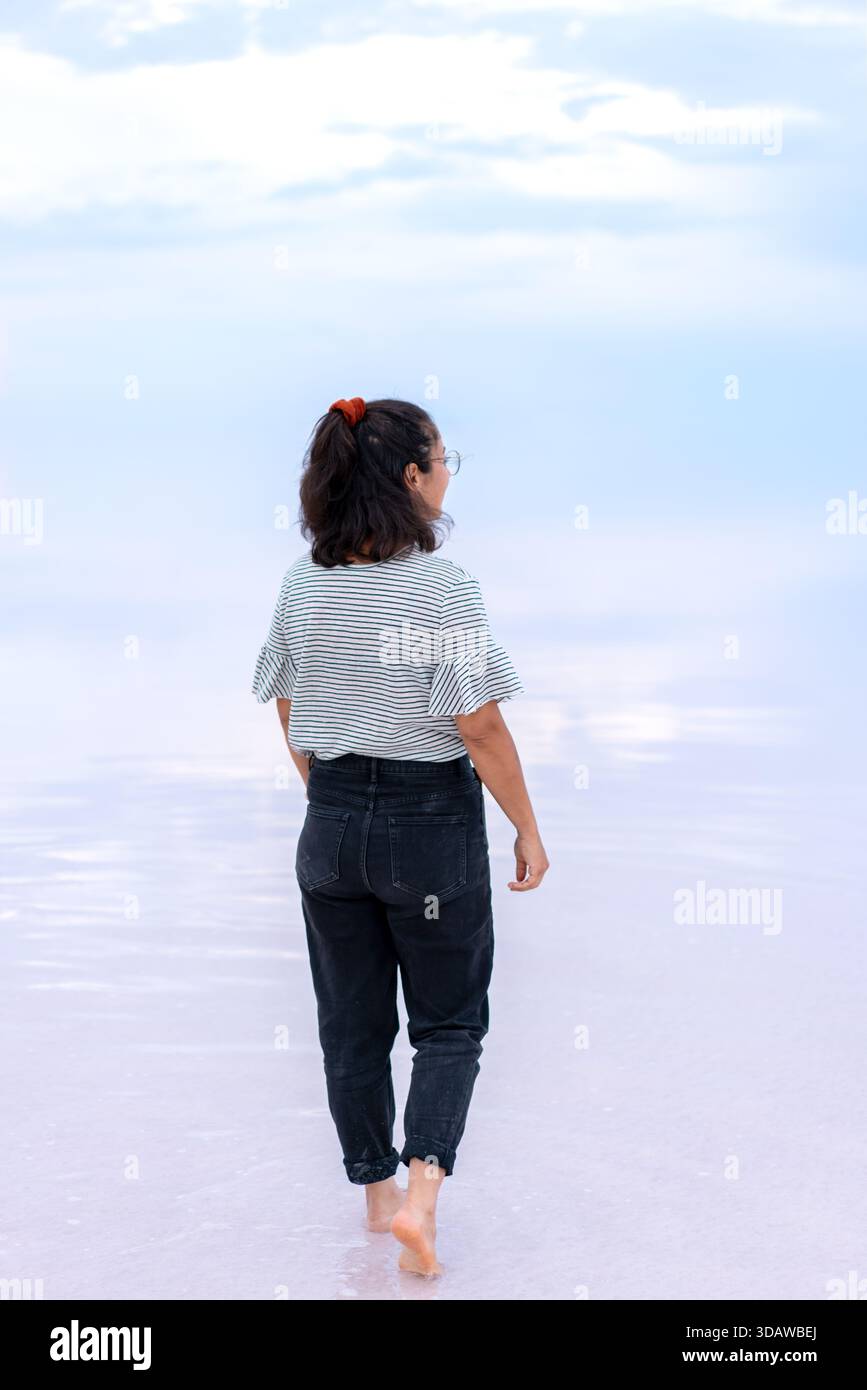 Una donna cammina a piedi nudi sul tranquillo lago salato di Tuz, creando uno specchio perfetto sulla superficie vetrata. L'ambiente tranquillo offre un ambiente rilassante Foto Stock