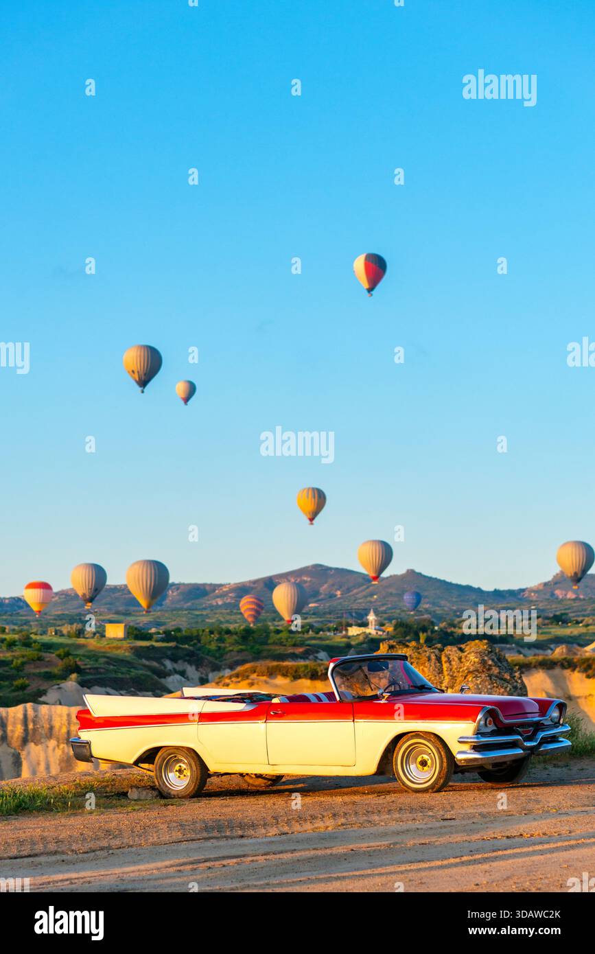 Una classica auto a noleggio decappottabile parcheggiata in Cappadocia all'alba con vivaci palloni ad aria calda che adornano il cielo, creando una scena pittoresca per il tour Foto Stock