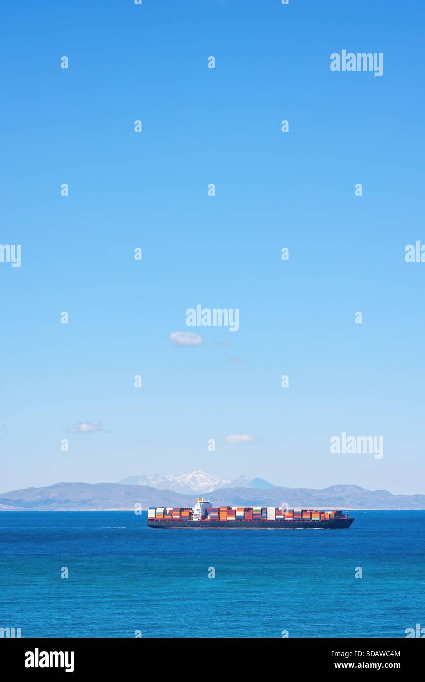 Una colorata nave da carico naviga nelle acque blu incontaminate con uno sfondo mozzafiato di montagne al largo della costa della Turchia, mostrando il commercio globale e il mare Foto Stock
