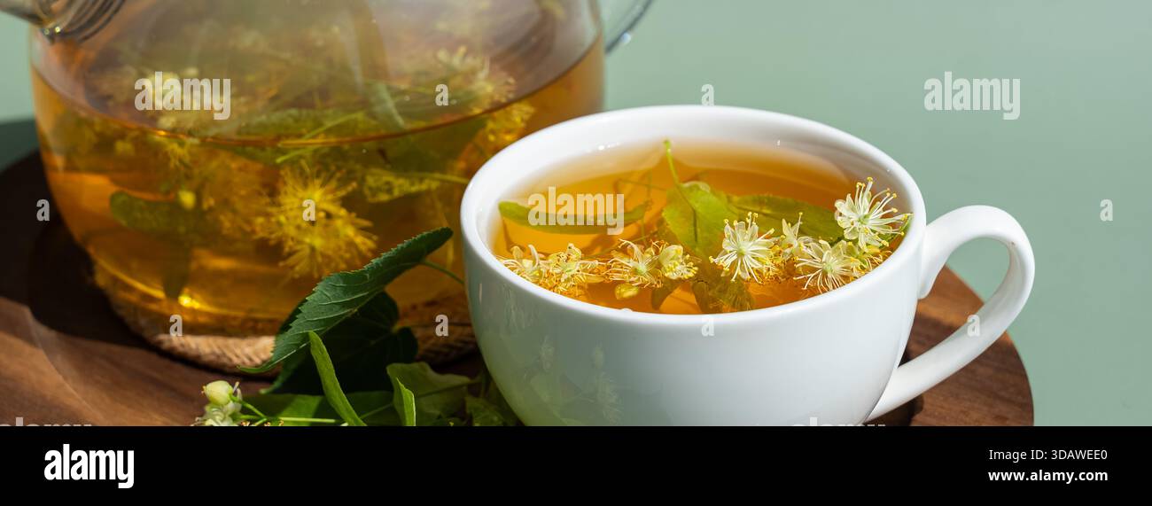 Una rilassante tazza di tiglio alle erbe con fiori freschi di tiglio che galleggiano in una tazza bianca. Una teiera trasparente con più infusione di tiglio è su un legno Foto Stock