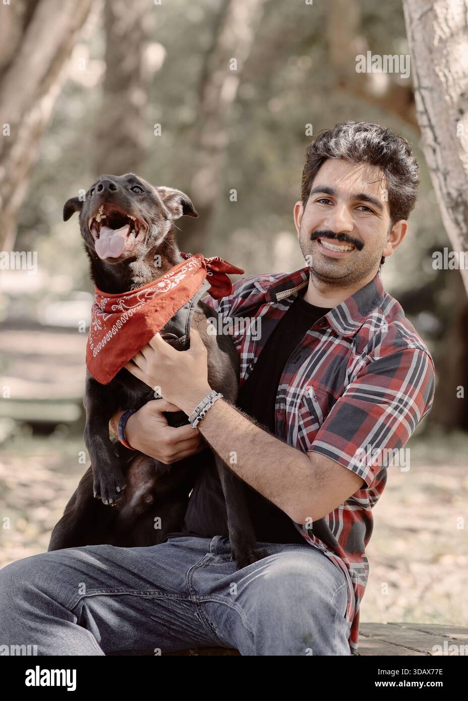 Momenti energetici con uomo e cane nel parco alberato. Cane nero con bandana rossa abbaiano o pantaloni. Il proprietario sorride alla telecamera. Luce naturale, stile di vita e animali domestici Foto Stock