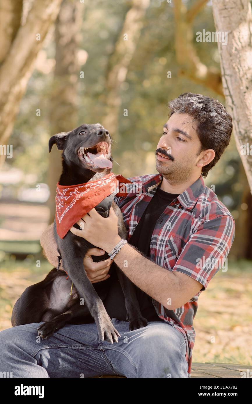 Uomo e cane si rilassano nel parco alberato. Cane nero con pantaloni bandana rossi felicemente. Il proprietario sorride alla telecamera. Luce naturale, legame emotivo, stile di vita e animali domestici Foto Stock