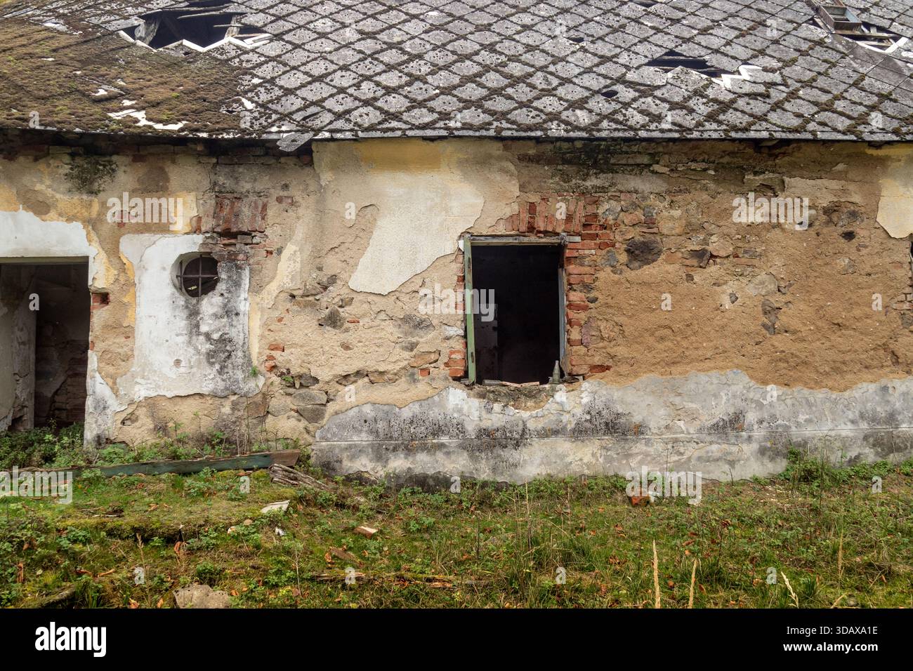 Facciata di una casa rurale abbandonata con intonaco danneggiato, finestre rotte e tetto deteriorato Foto Stock