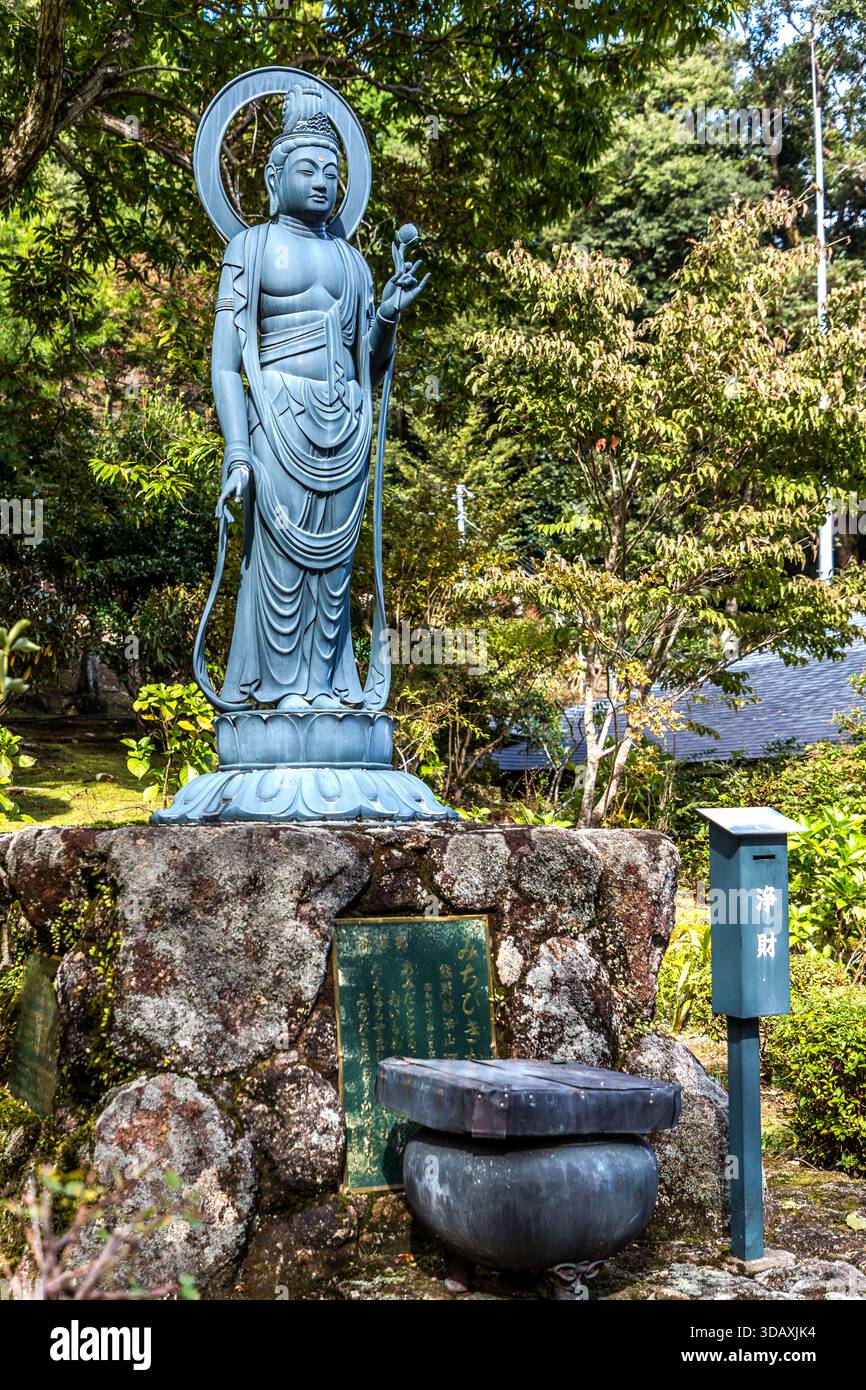 Una statua di Buddha si erge su un piedistallo di pietra nel giardino del tempio Myōhōzan Amida-ji vicino a Nachikatsuura, prefettura di Wakayama, Giappone. Una scatola delle offerte e una targa con iscrizioni si trovano davanti ad essa. ## Batch 27 Foto Stock