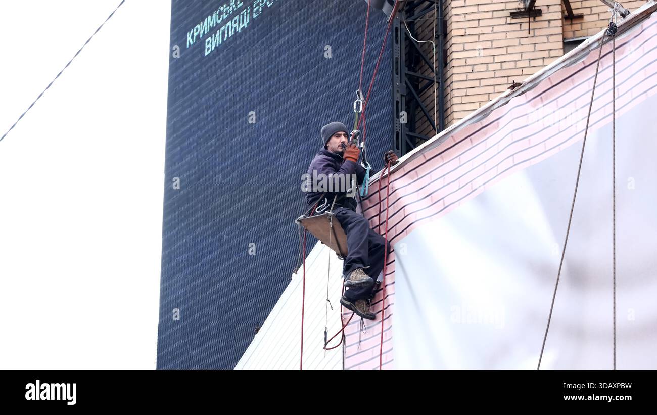 Un operaio qualificato pende da una sporgenza, fissando l'esterno di un alto edificio in centro. Kiev, Ucraina. 12 dicembre 2025. Foto Stock