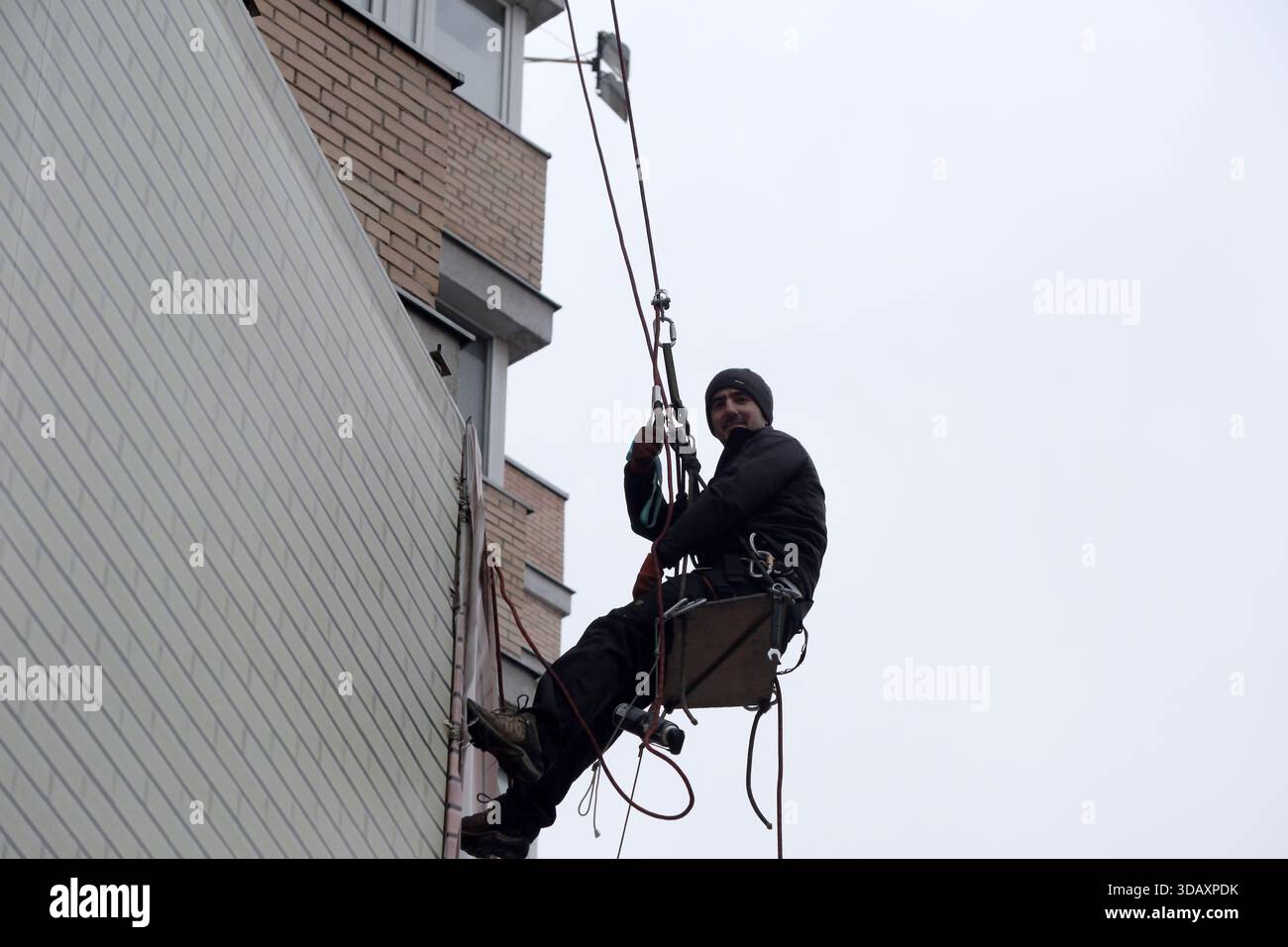 Un lavoratore in un imbracatura esegue la manutenzione su un edificio alto, sospeso a mezz'aria. Kiev, Ucraina. 12 dicembre 2025. Foto Stock