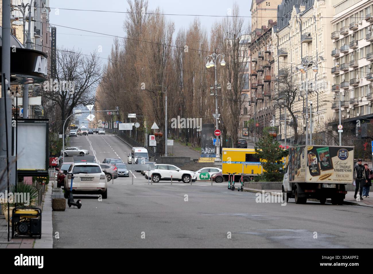 Le auto attraversano una strada vivace delimitata da alti alberi sotto un cielo grigio. Kiev, Ucraina. 12 dicembre 2025. Foto Stock