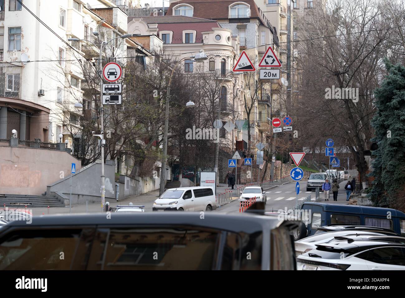 Le auto sono parcheggiate lungo una strada stretta con persone che camminano e segnali stradali visibili. Kiev, Ucraina. 12 dicembre 2025. Foto Stock