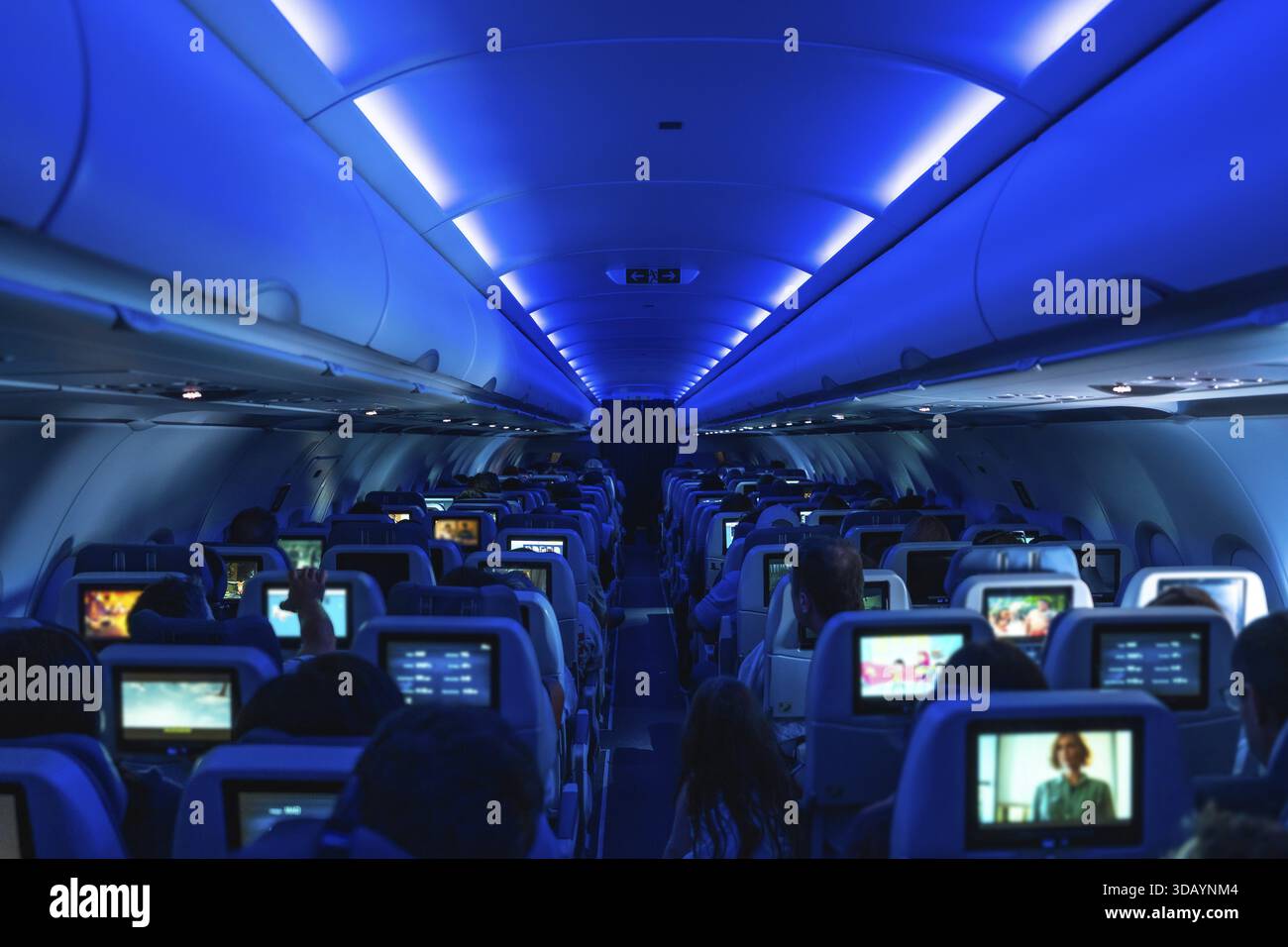 I passeggeri si godono l'intrattenimento durante il volo mentre guardano film su schermi personali, circondati da un'atmosfera accogliente sotto l'illuminazione blu Foto Stock