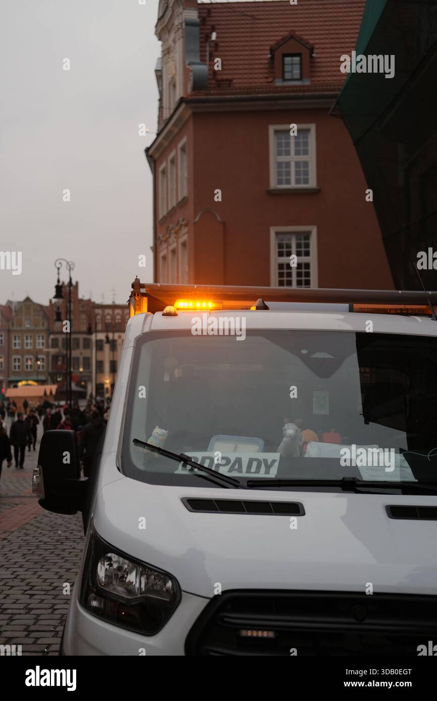 Vista frontale del furgone multiuso bianco con spie ambra accese sul tetto, parcheggiato su una strada acciottolata, edifici storici sullo sfondo. Foto Stock