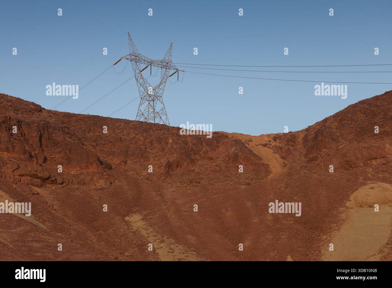 Un pilone in acciaio si erge contro un cielo blu limpido, torreggiante su colline ripide, aride e rossastre, mostrando l'industria che incontra la natura remota. Foto Stock