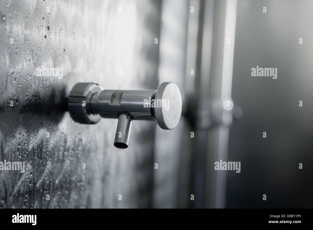 Primo piano di una valvola del serbatoio di fermentazione del vino in acciaio inossidabile, con gocce di umidità sulla superficie, che evocano un'atmosfera industriale ma pulita dell'azienda vinicola. Foto Stock