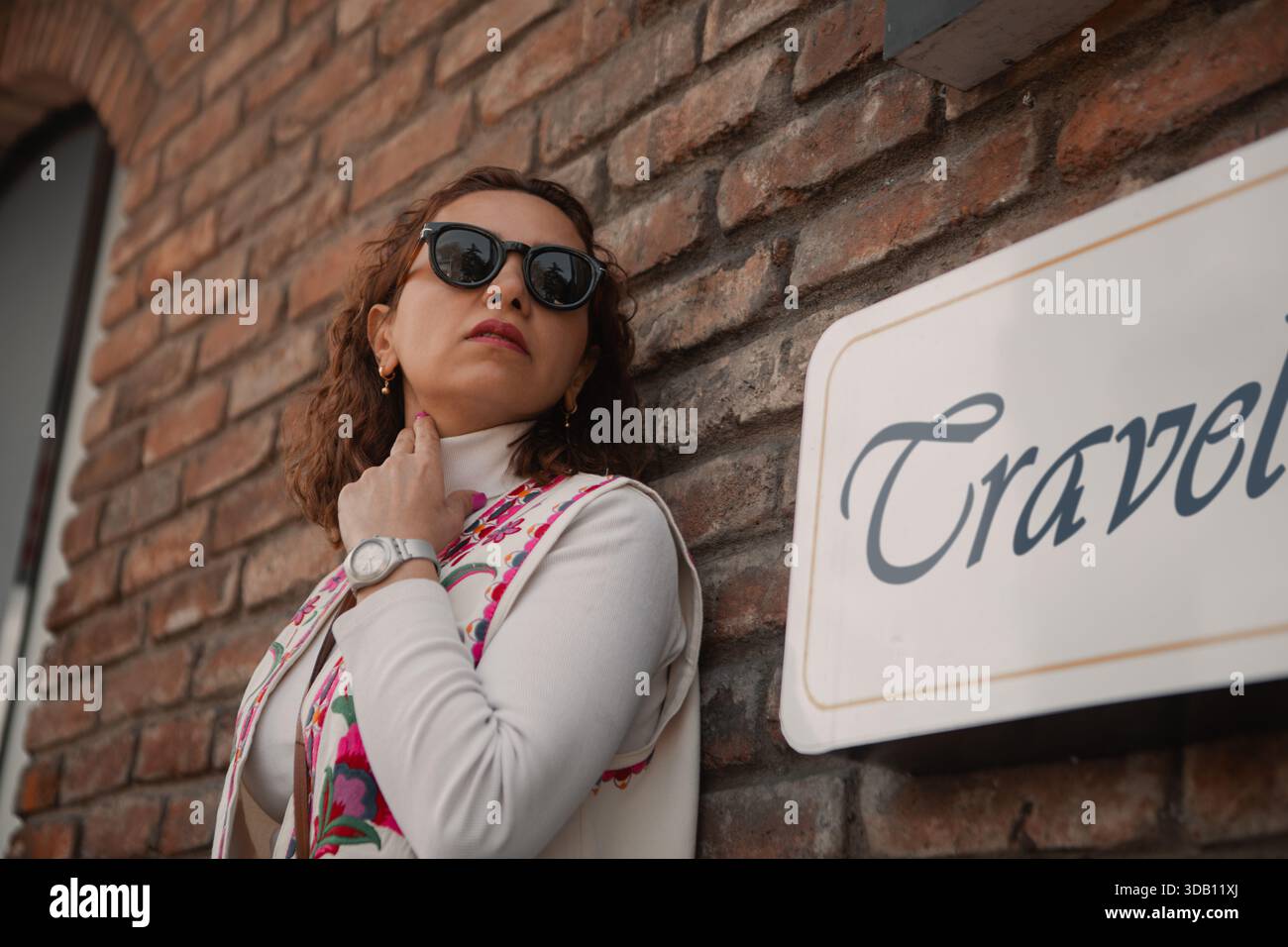 Una donna elegante con occhiali da sole si appoggia con sicurezza contro un muro di mattoni accanto a un cartello che legge Foto Stock