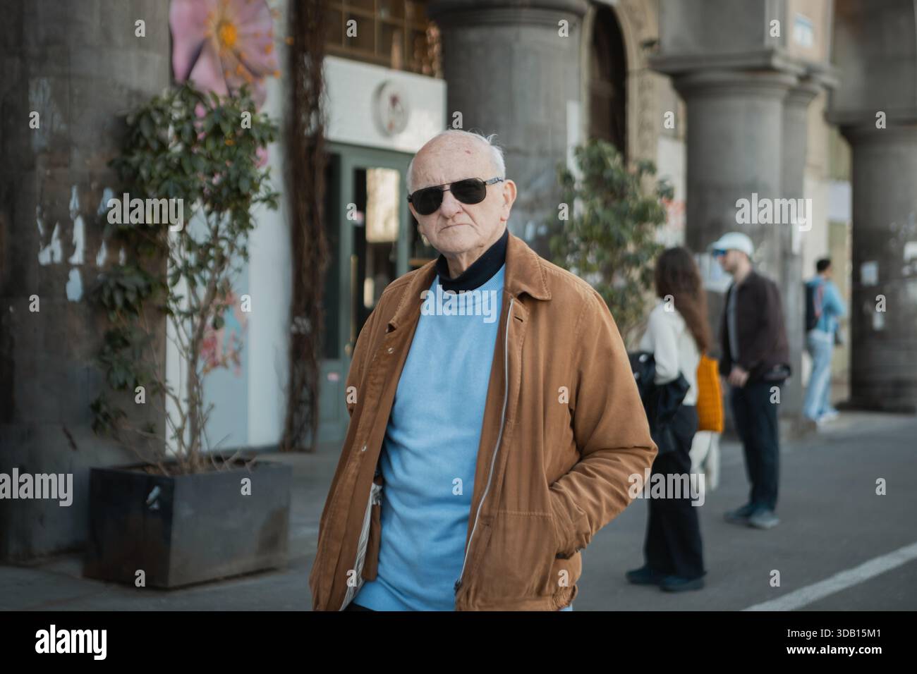 Tbilisi, Georgia, 27 marzo 2025: Un uomo più anziano con giacca marrone e occhiali da sole si trova fuori da un edificio colonnato, con due figure sfocate in... Foto Stock