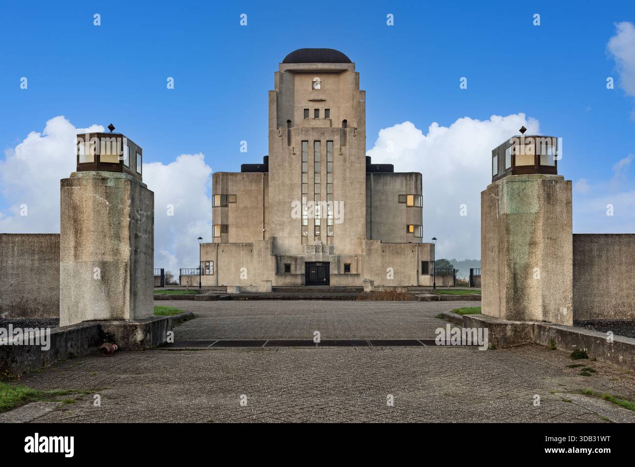 Vista frontale simmetrica del monumentale edificio radio Kootwijk, perfettamente incorniciato da pilastri d'ingresso sotto un cielo blu con nuvole, in mostra Art De Foto Stock