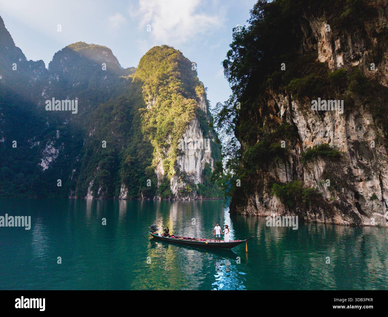 Una coppia di fronte a una barca a coda lunga all'alba sul lago Cheow LAN a Khao Sok, Thailandia. Le acque color smeraldo sono circondate da torreggianti scogliere calcaree, che offrono uno sfondo perfetto per le avventure Foto Stock