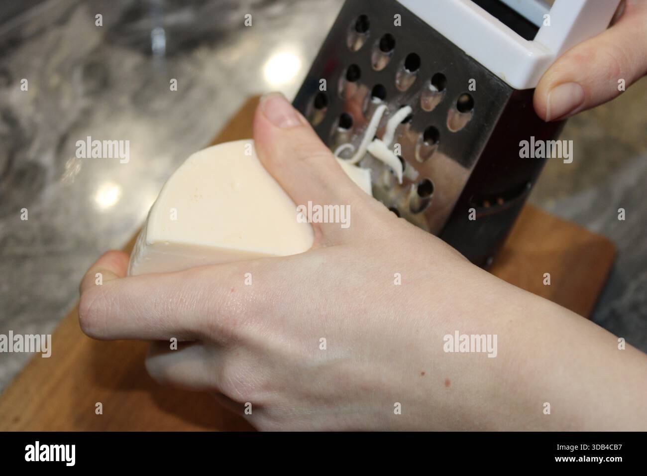 Una persona sta grattugiando un blocco di formaggio con una grattugia metallica mentre si trova in piedi su un tagliere di legno. La cucina ha un'atmosfera luminosa e materiali Foto Stock