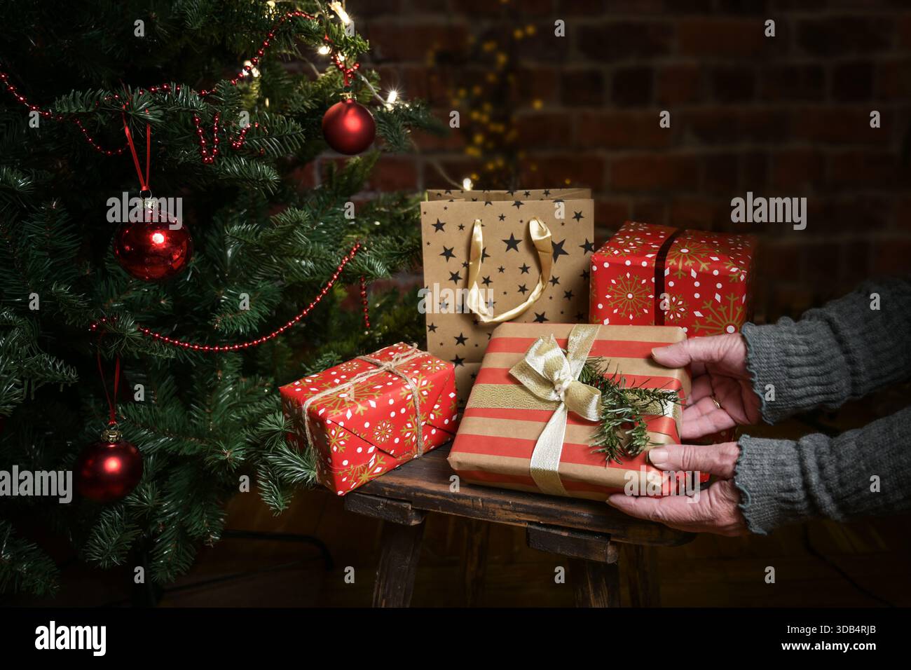 Mani di una donna anziana che mette regali avvolti sotto un albero di Natale decorato contro un muro di mattoni scuri, concetto per donare con amore e donazione co Foto Stock