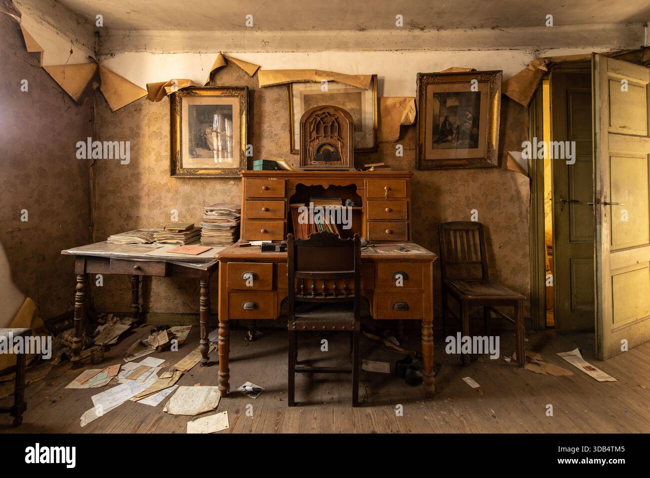 Studio vintage abbandonato con scrivania in legno, radio antica, carta da parati sbucciata e carte sparsi in una stanza piena di luce abbandonata Foto Stock