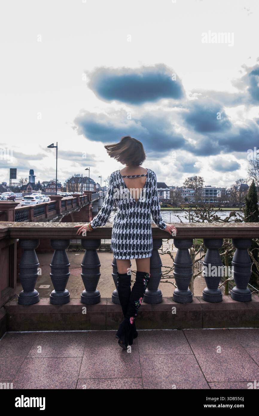Una donna vista da dietro si erge sul ponte pedonale sul fiume meno a Francoforte, indossando un elegante abito monopezzo in bianco e nero. Foto Stock