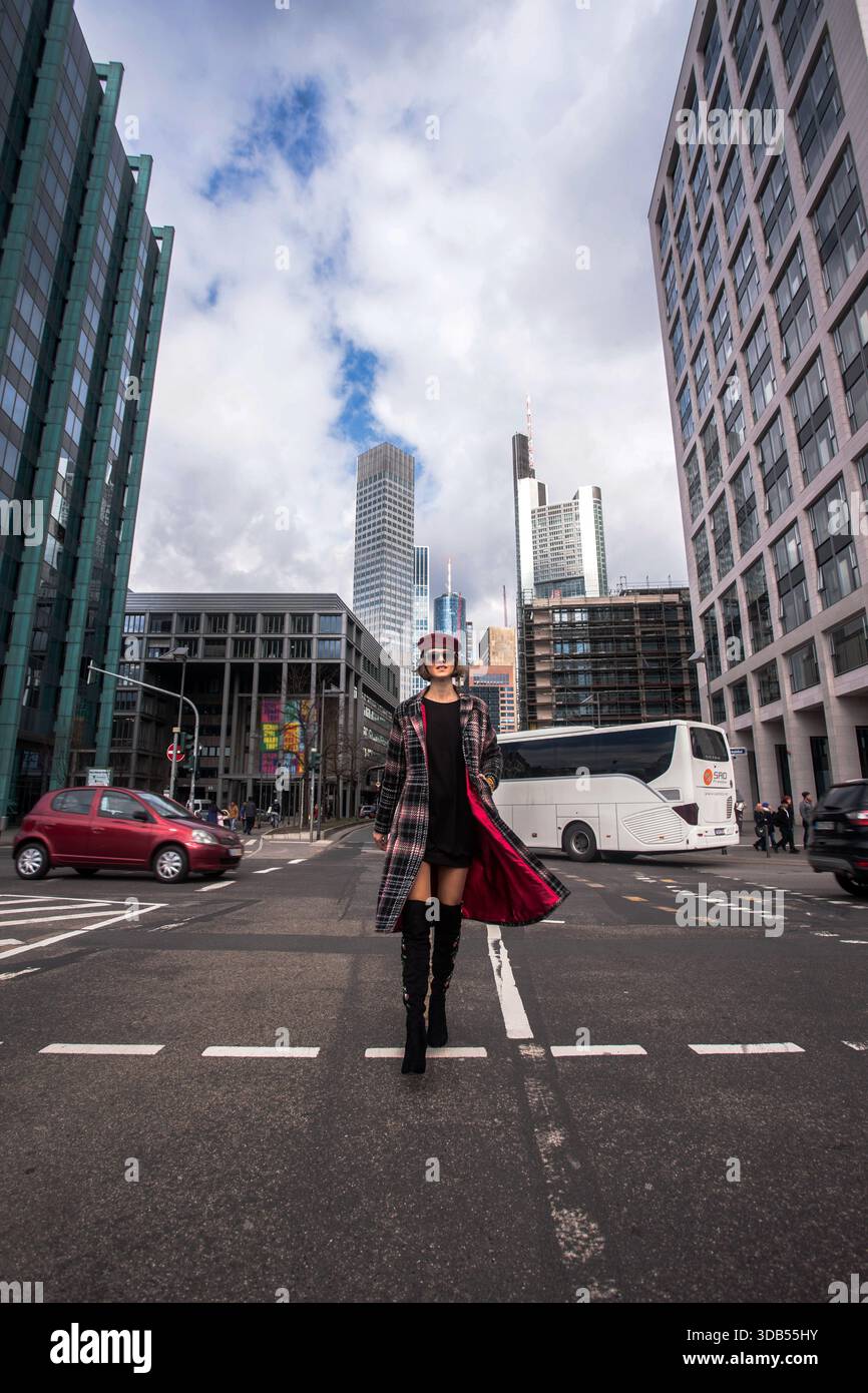 Una donna alla moda e sicura di sé che indossa stivali alti, un cappotto lungo e un cappello militare rosso cammina attraverso una strada della città con i moderni grattacieli. Foto Stock