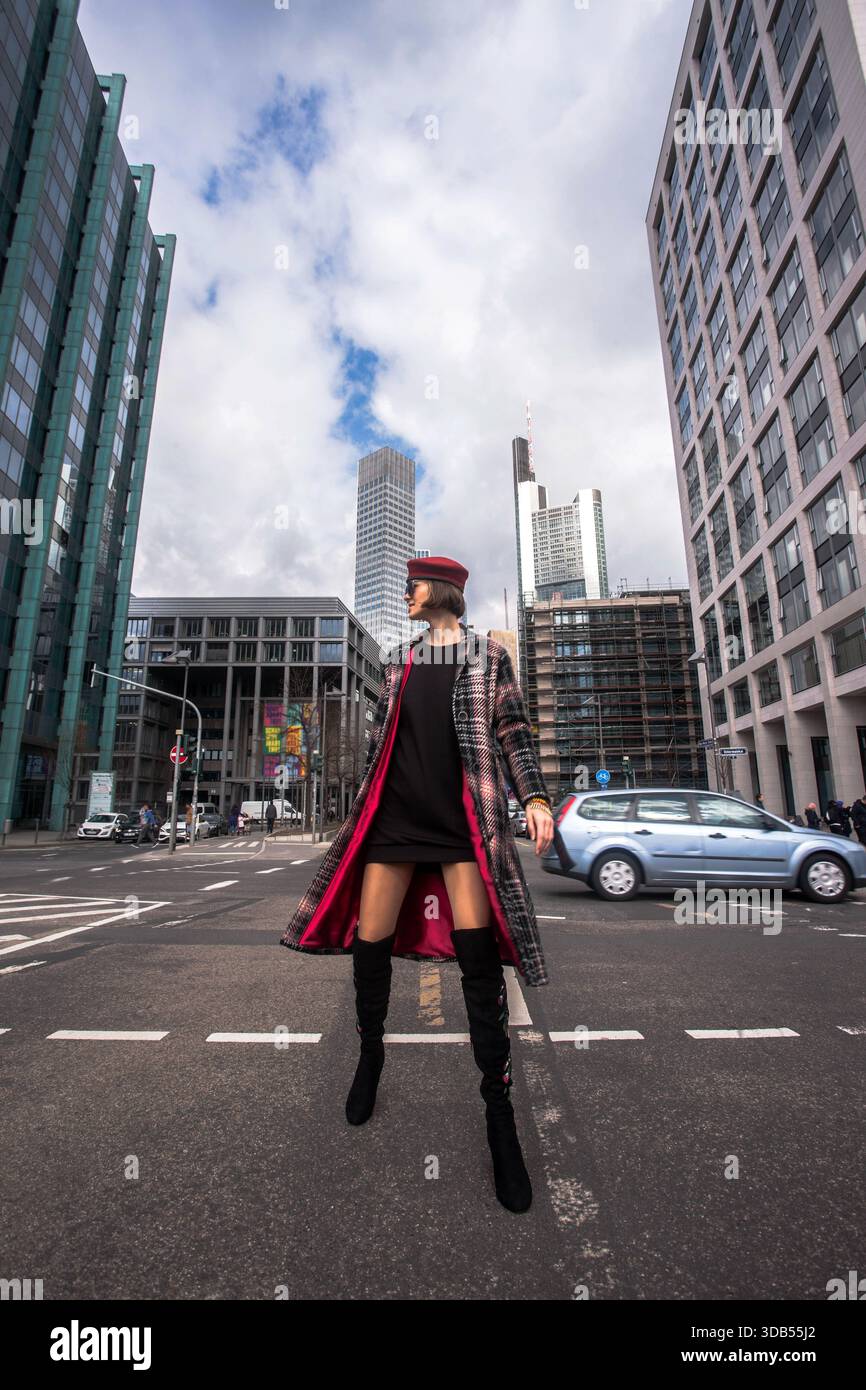 Una donna alla moda e sicura di sé che indossa stivali alti, un cappotto lungo e un cappello militare rosso cammina attraverso una strada della città con i moderni grattacieli. Foto Stock