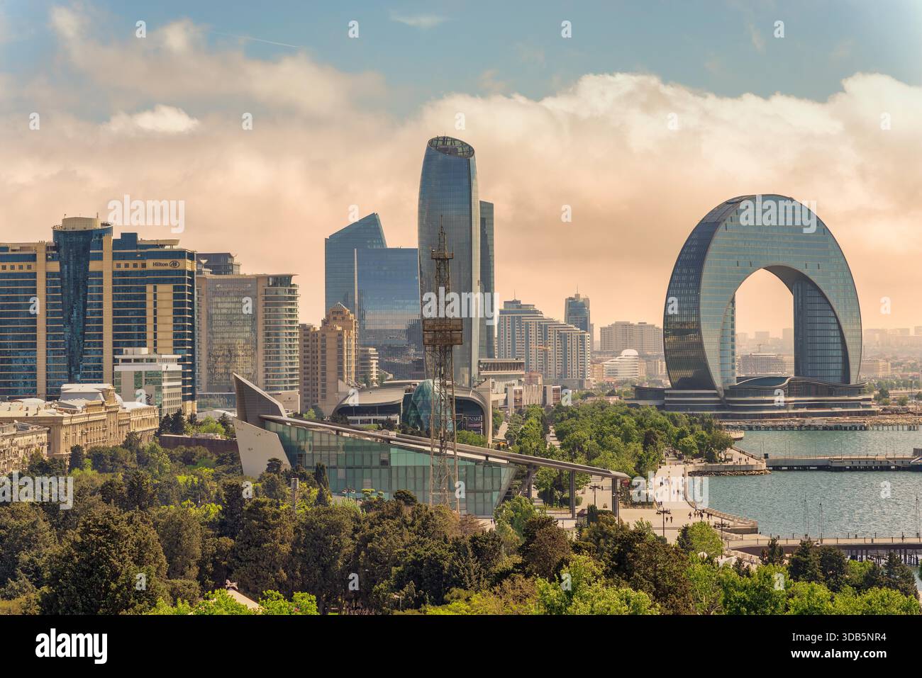Baku, Azerbaigian - 10 maggio 2024: Paesaggio urbano caratterizzato dall'iconico Crescent Hotel, dalla SOCAR Tower e dall'Hilton hotel. Architettura moderna, parco verde e lungomare sotto un cielo nuvoloso Foto Stock