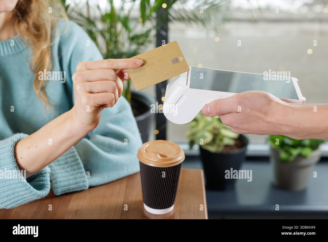Donna che tocca la carta di credito su un lettore di carte portatile per il pagamento contactless NFC al tavolo del caffè con caffè da asporto e piante Foto Stock
