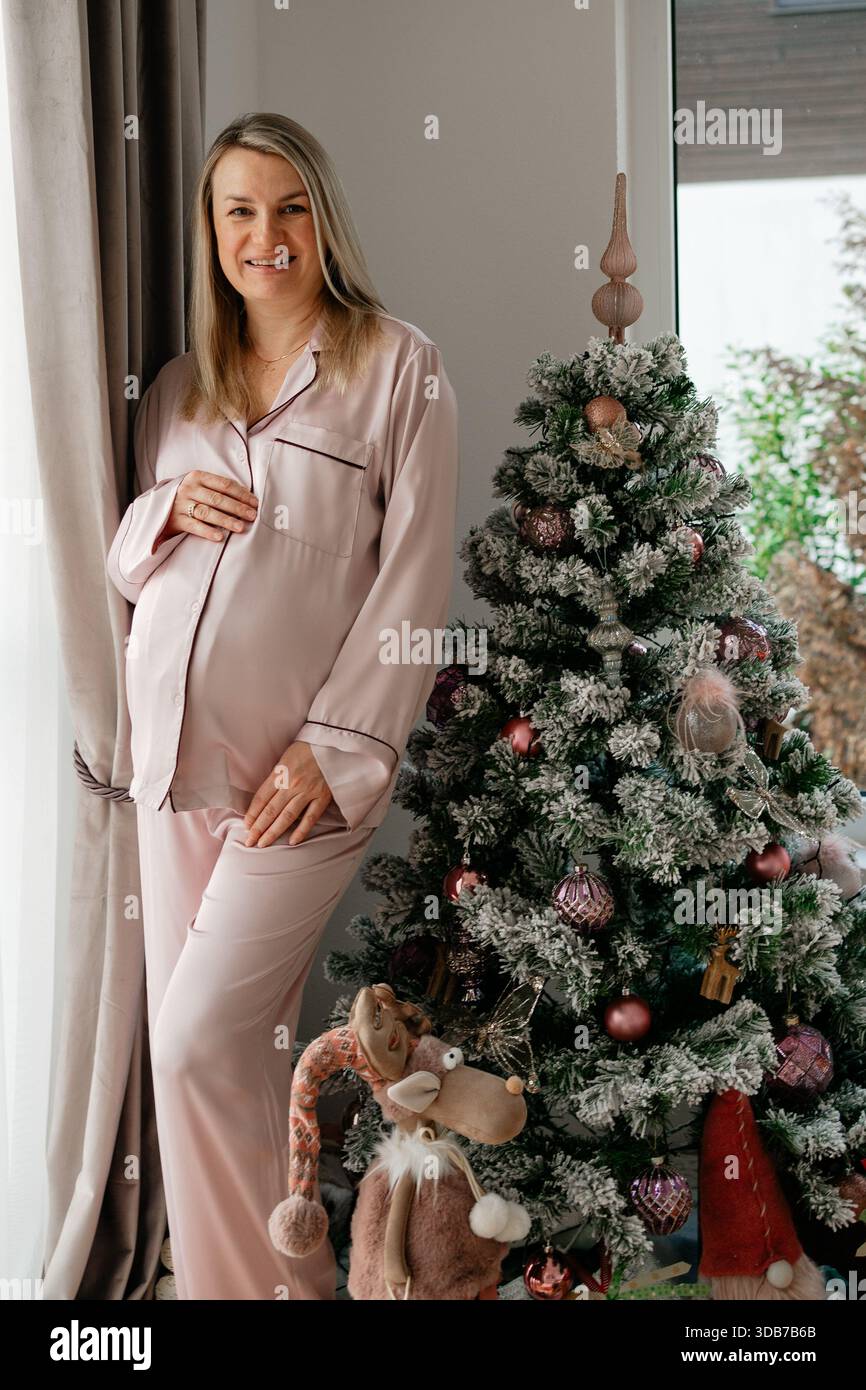 Una donna incinta a 36 settimane si trova accanto a un albero di Natale decorato in un luminoso interno domestico. Luce soffusa del giorno, abbigliamento da salotto pastello e dettagli festivi Foto Stock