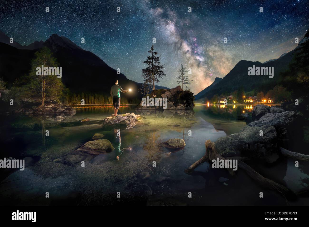 Magica scena blu in un lago di notte, con un uomo in piedi sotto la via Lattea su una roccia e che tiene una luce sull'acqua calma Foto Stock