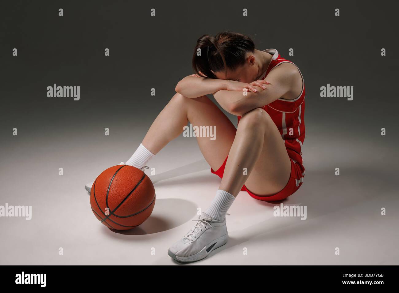Stanca giocatrice di basket si siede con la testa in giù, mostrando stress ed esaurimento emotivo dopo la partita in uno studio. Foto Stock