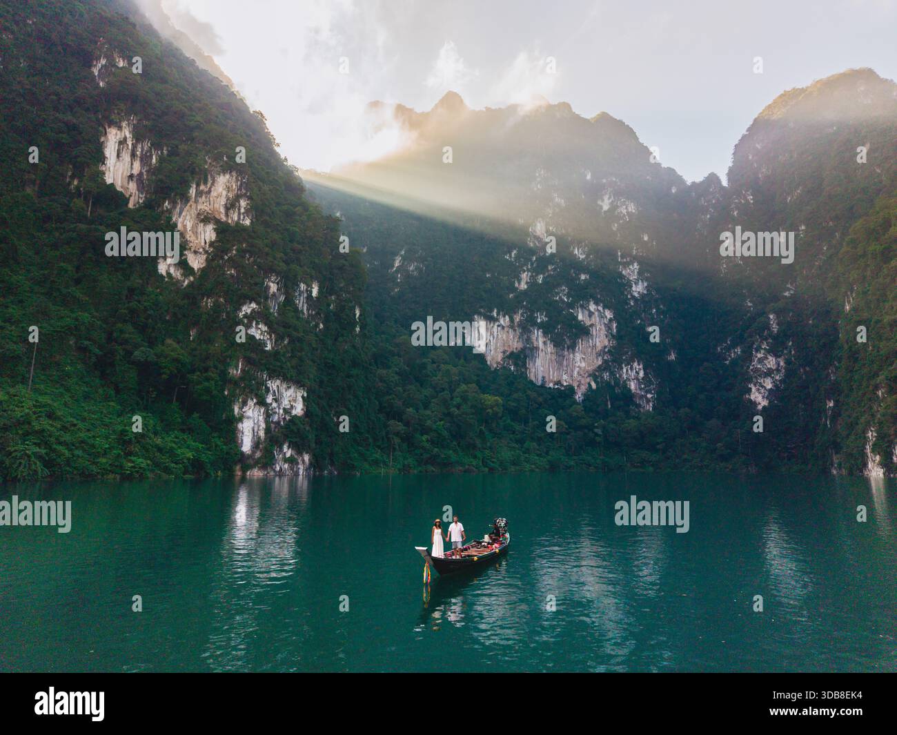 Osserva le tranquille acque del lago Cheow LAN a Khao Sok, Thailandia, durante l'alba. Una coppia su una barca a coda lunga scivola senza intoppi attraverso il lago mentre il sole getta raggi dorati sulle scogliere torreggianti. Foto Stock