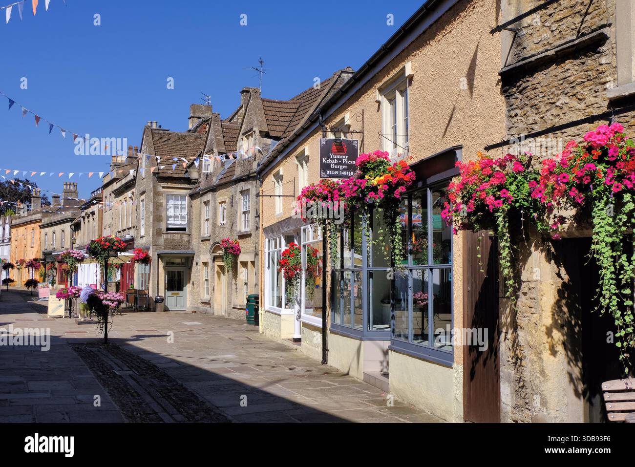 Corsham: Edifici d'epoca, negozi, caffetterie, leoni dipinti e fiori in High Street con cielo blu, Corsham, Wiltshire Foto Stock