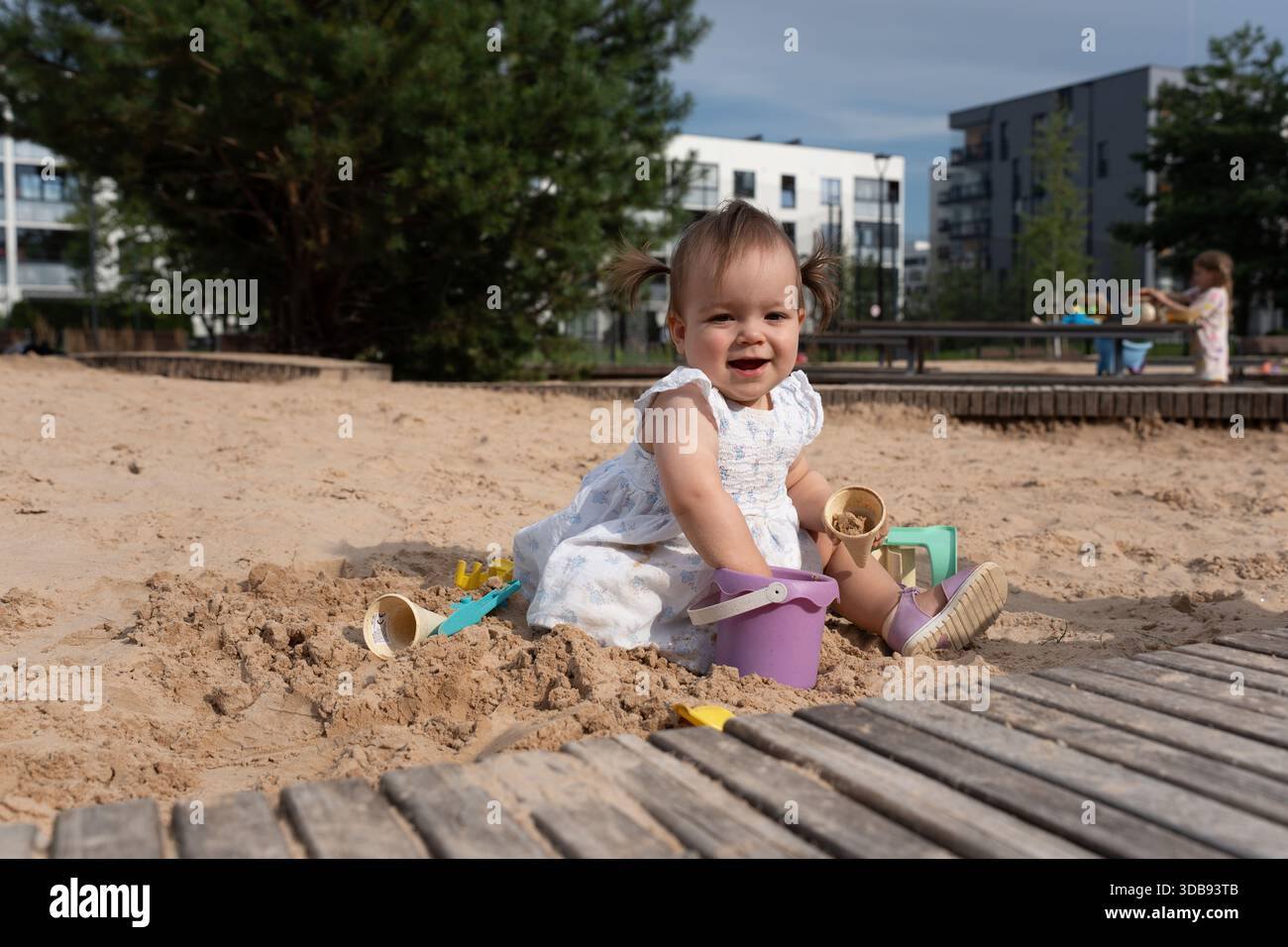 un bambino allegro che gioca in una sandbox urbana illuminata dal sole con giocattoli colorati ed edifici residenziali sullo sfondo in una giornata limpida, curiosità, spensieratezza, Foto Stock
