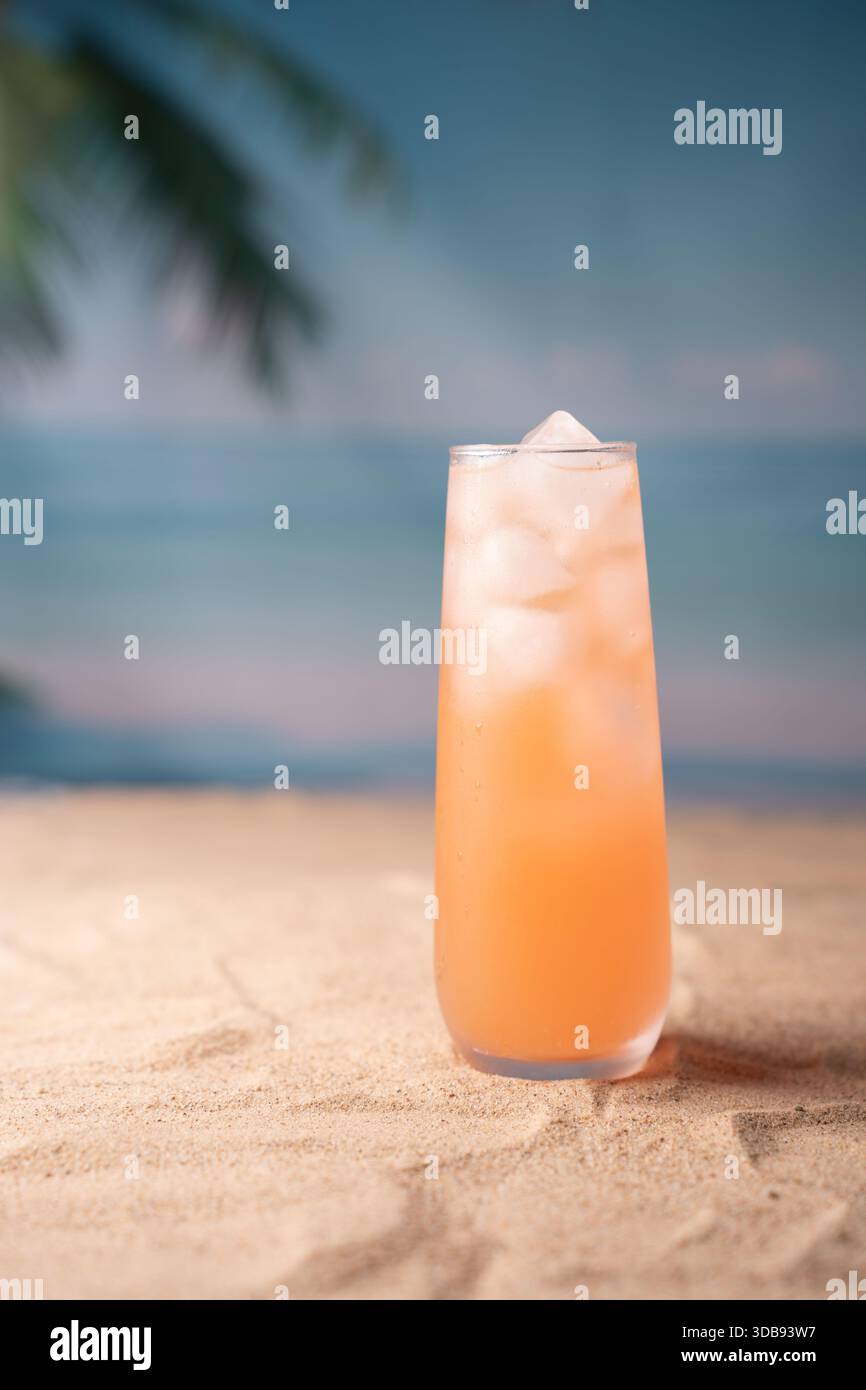 rinfrescante cocktail di agrumi rosa in alto vetro con ghiaccio sulla spiaggia di sabbia sotto il cielo azzurro e rilassante sullo sfondo dell'oceano, evocando idilliaci vacanze tropicali Foto Stock