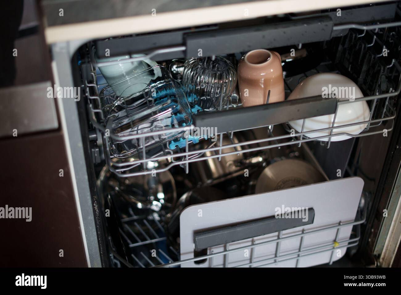 lavastoviglie aperta con un assortimento di piatti, bicchieri e utensili, che mostrano una miscela di materiali e colori tenui in un ambiente di cucina casalinga Foto Stock