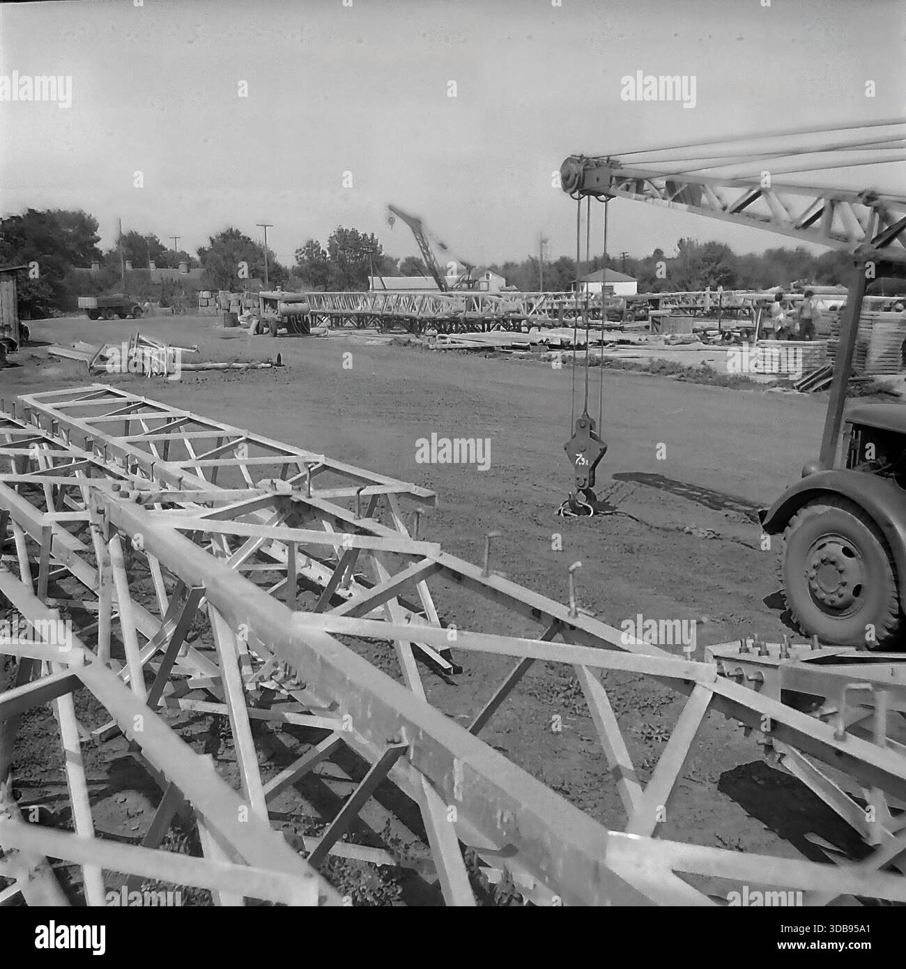 1970 anni URSS. Regione di Donetsk. Costruzione di linee elettriche strategiche ad alta tensione da 750 kV. Vista del cantiere di assemblaggio in cui vengono preparate massicce sezioni a reticolo in acciaio delle torri di trasmissione. Un pesante gancio per gru pende sopra il sito e una gru per camion sovietica d'epoca è visibile sulla destra. Sullo sfondo, le strutture metalliche sono impilate e pronte per il trasporto. Un documentario storico girato che illustra la scala industriale dello sviluppo delle infrastrutture energetiche nella pacifica steppa del Donbas durante l'era sovietica. Foto Stock