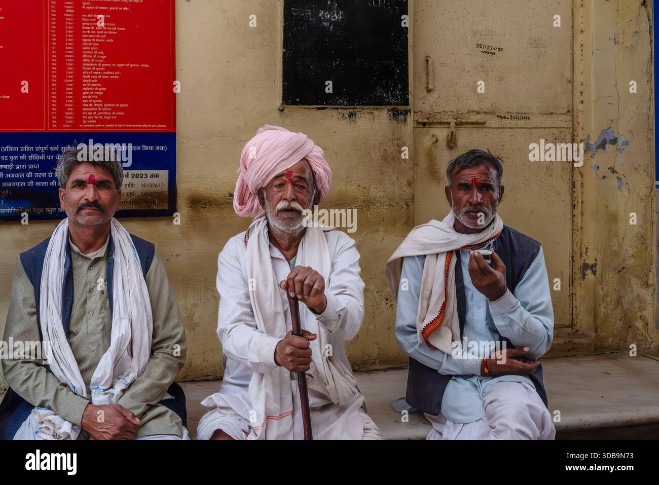 Tre anziani uomini indiani in abiti tradizionali siedono su una panchina in un'area pubblica in India, catturando la vita quotidiana e la cultura rurale. Foto Stock